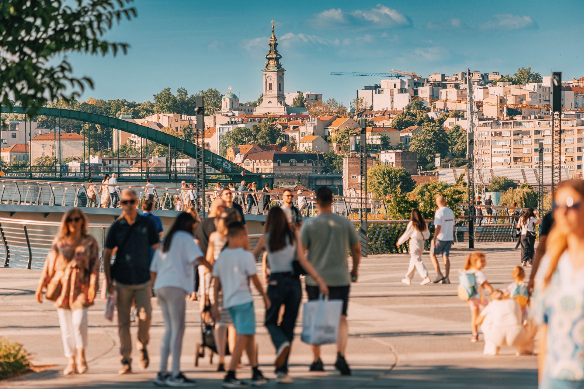 Belgrad’da Sava kıyısındaki yürüyüş yolunda gezinen insanlar, arkada eski şehir ve bir kilise kulesi